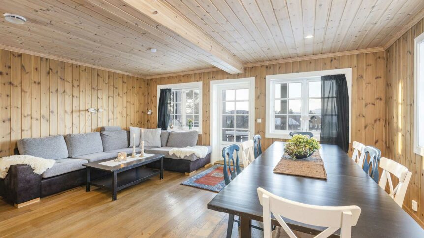 Skandinavisk stue med trepaneler, grå sofa, spisebord og store vinduer som slipper inn naturlig lys.
