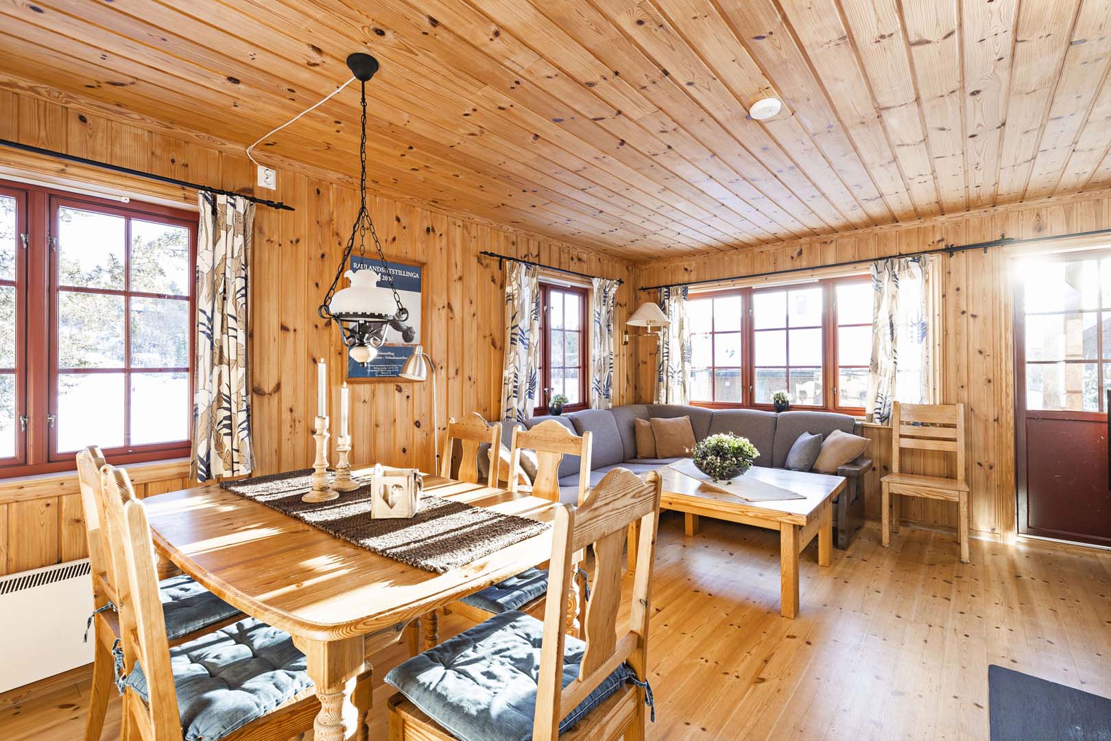 Lyst tømmerstue med spisebord, sofa og store vinduer som slipper inn naturlig lys. Koselig norsk interiør.