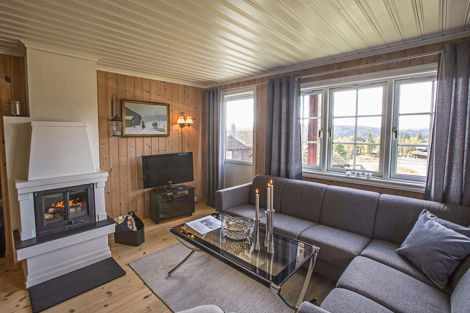 Koselig stue med moderne peis, TV og hjørnesofa, omgitt av store vinduer med utsikt over naturen.