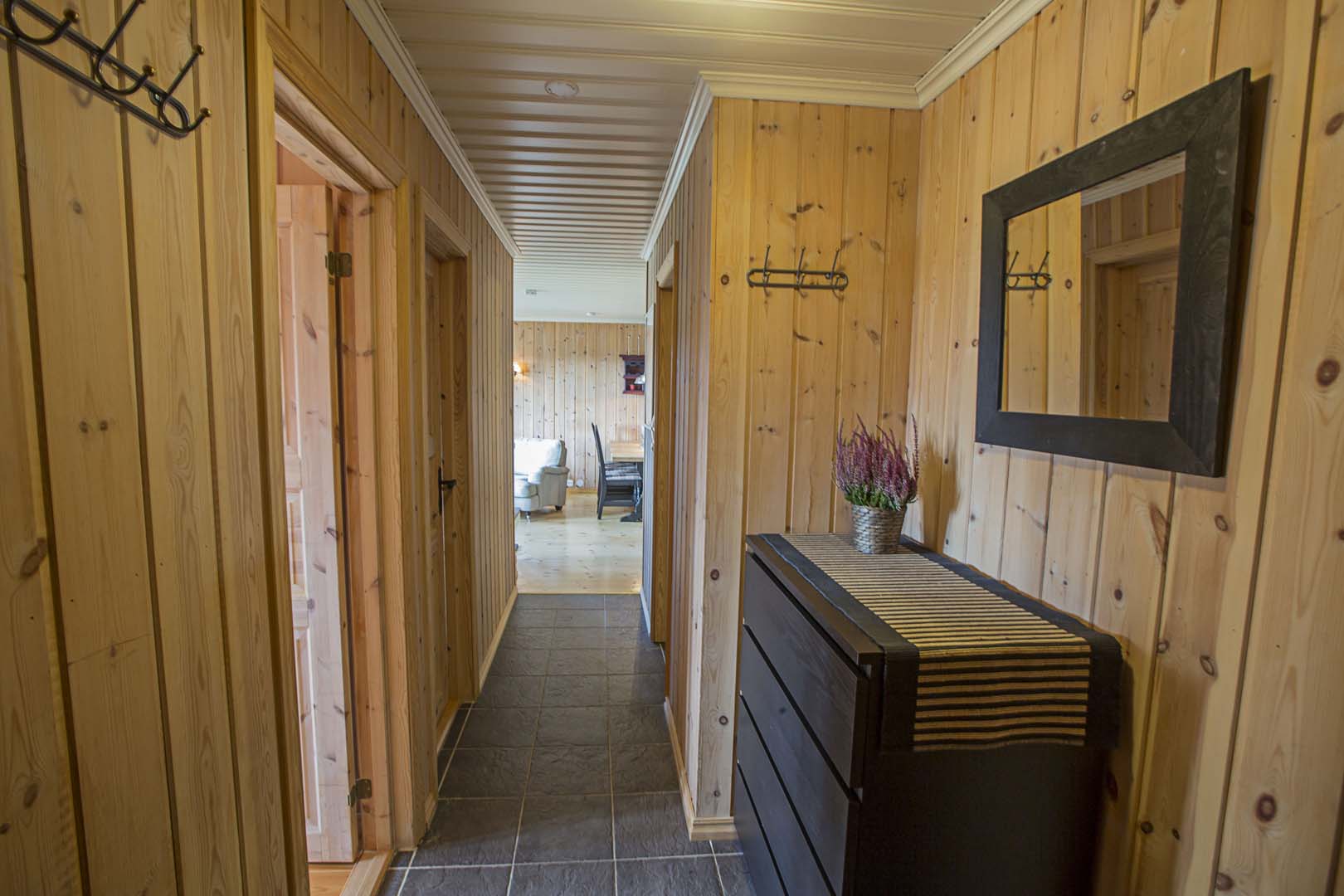 Gang i trehytte med speil, kommode og utsikt til stue. Hyttestil med paneler og flislagt gulv.