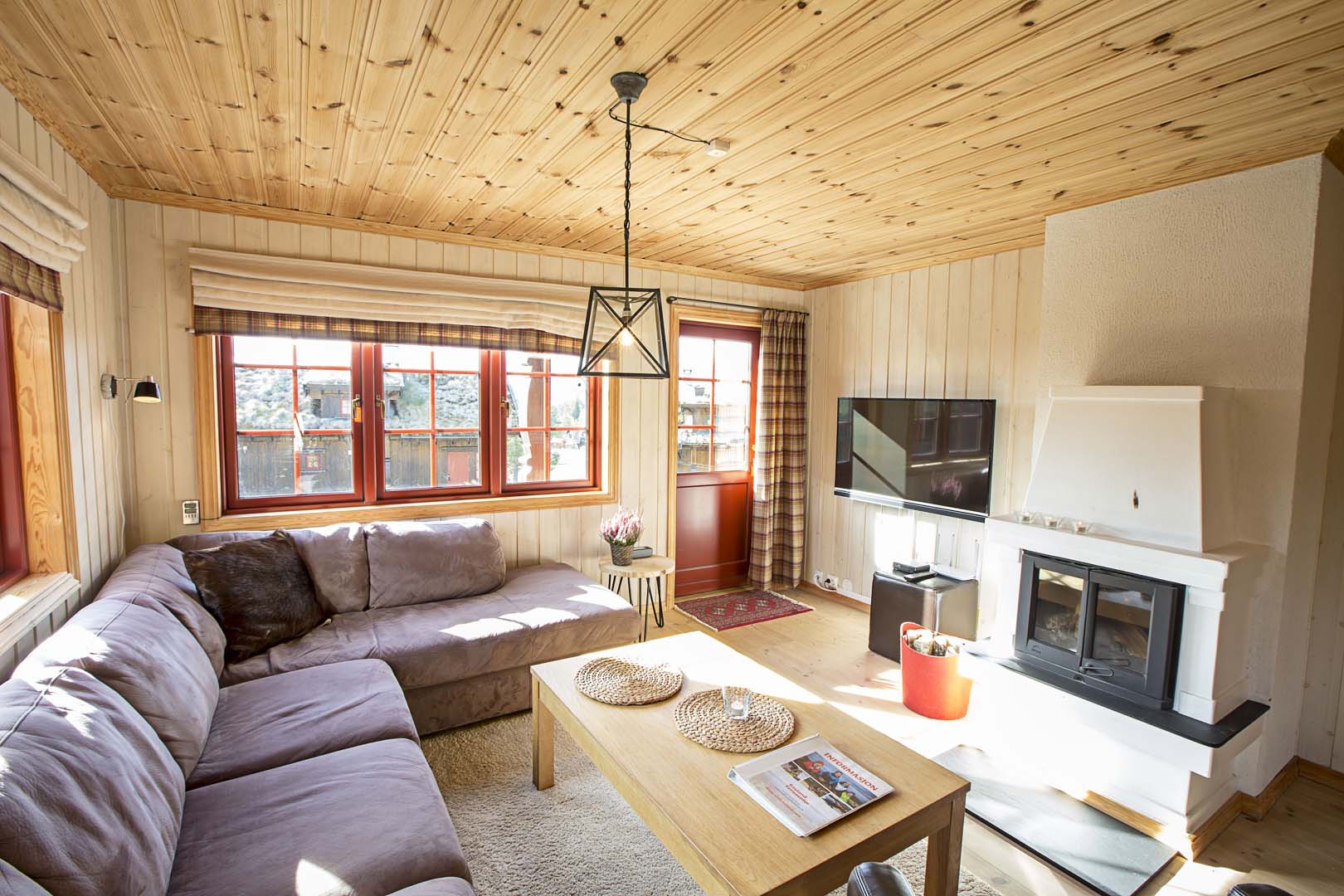 Koselig stue med hjørnesofa, peis, trepanel og store vinduer, ideelt for avslapning i et norsk fjellhytteinteriør.