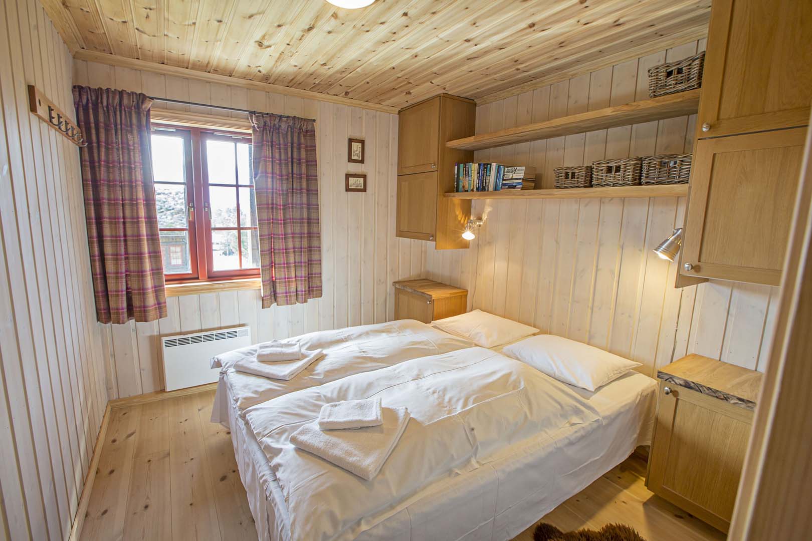 Koselig soverom i trehytte med dobbeltseng, vindu med rutete gardiner og hyller med bøker.