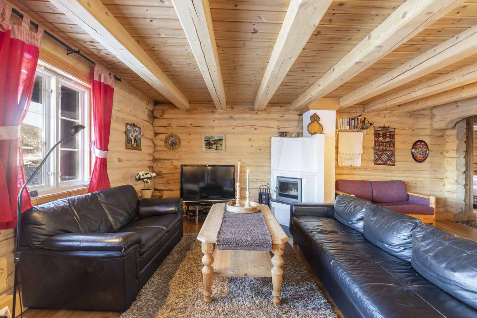 Stue i tømmerhytte med svarte sofaer, peis, rød gardiner og naturlig treinteriør. Koselig og tradisjonell atmosfære.