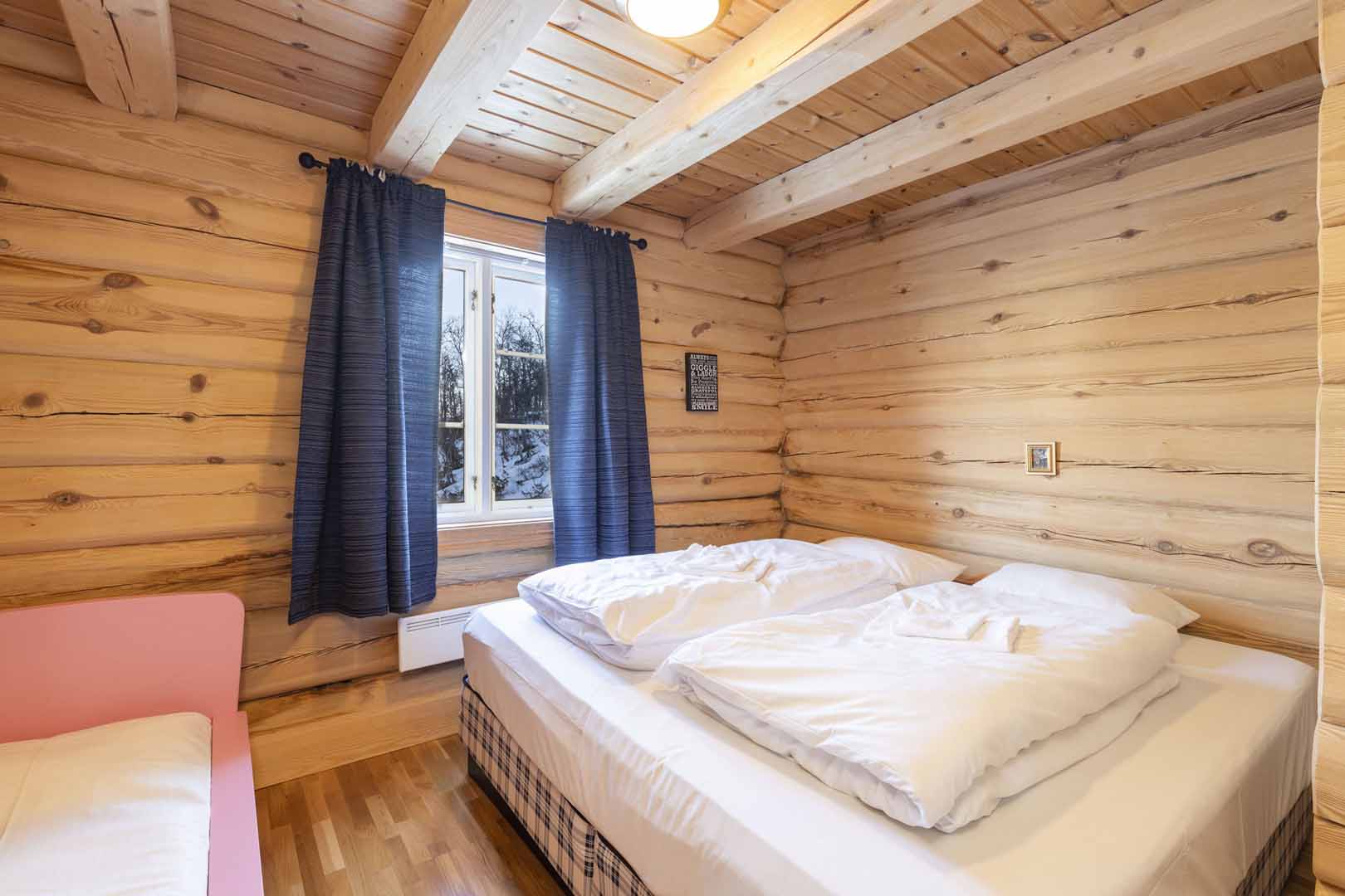 Soverom i norsk tømmerhytte med dobbeltseng, hvitt sengetøy og utsikt mot snødekte trær gjennom vinduet.