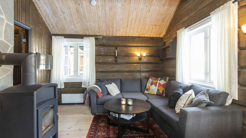 Koselig stue med grå sofa, peis, rustikke tømmervegger og fargerike puter i en norsk hytte.