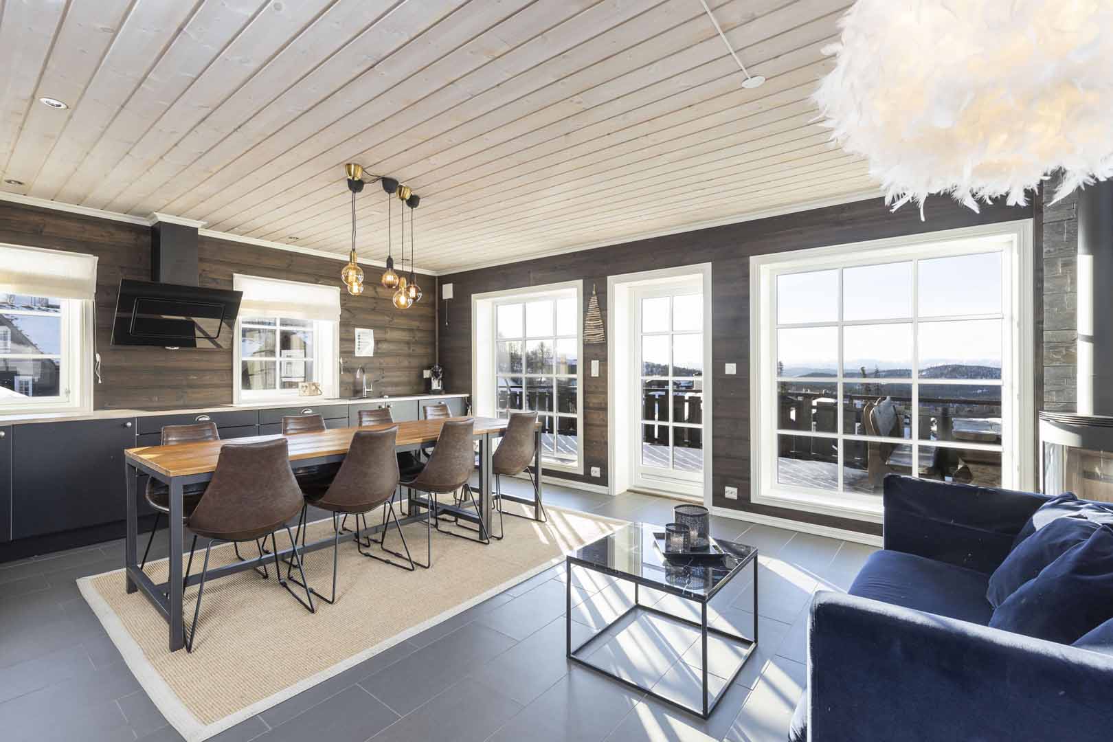 Moderne stue med spisebord, store vinduer og fjellutsikt. Elegant belysning og blå sofa i koselige omgivelser.