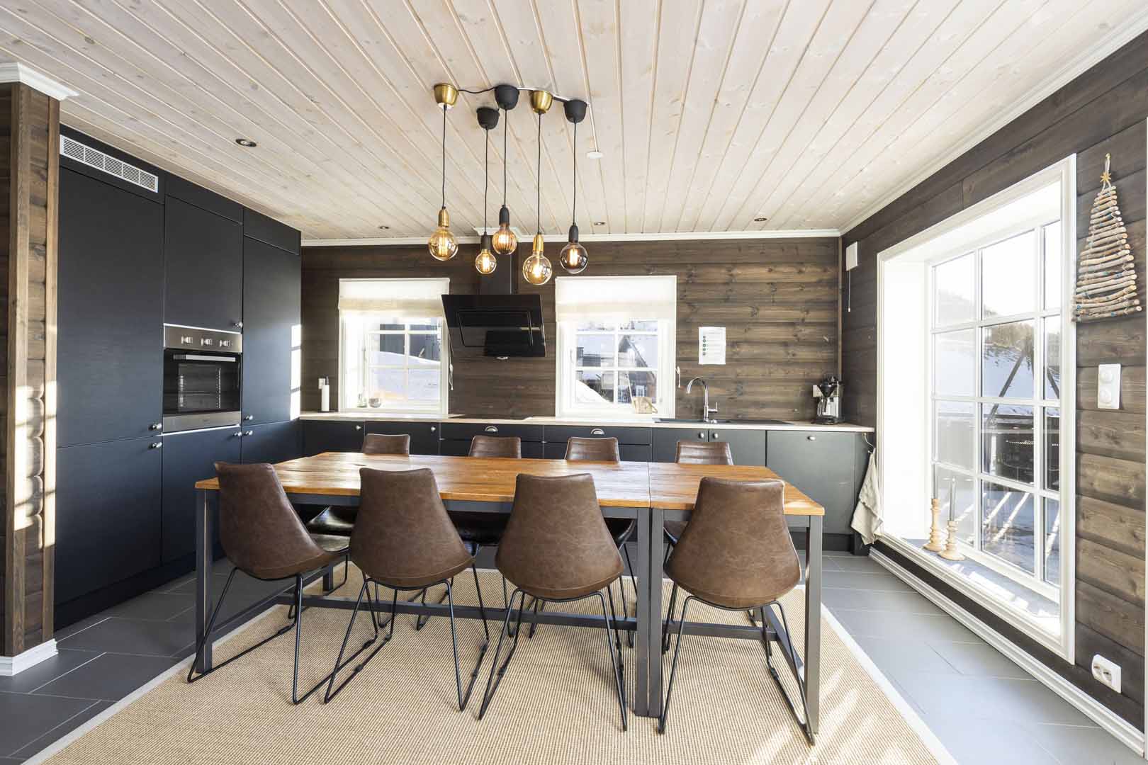 Moderne kjøkken med tre-bord, skinnstoler og hengelamper i rustikk stil. Store vinduer slipper inn naturlig lys.