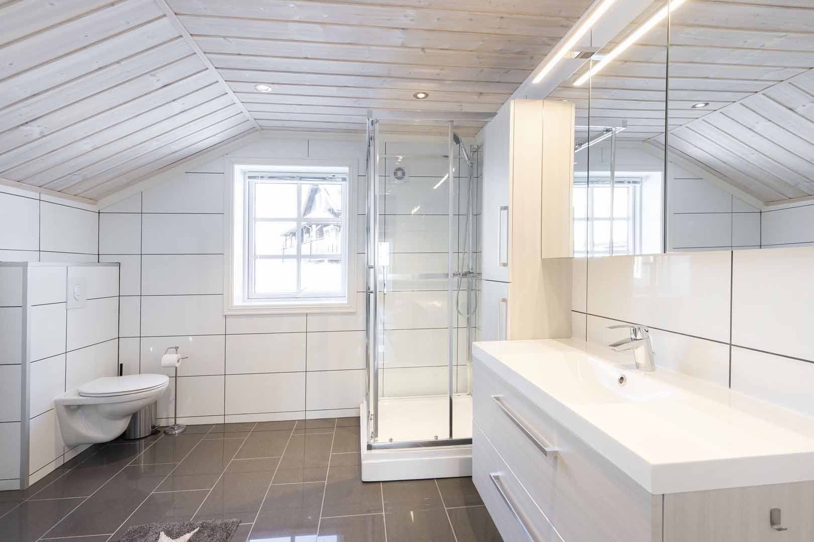 Moderne bad med flislagte vegger, dusjkabinett, toalett og speil skap under skråtak.