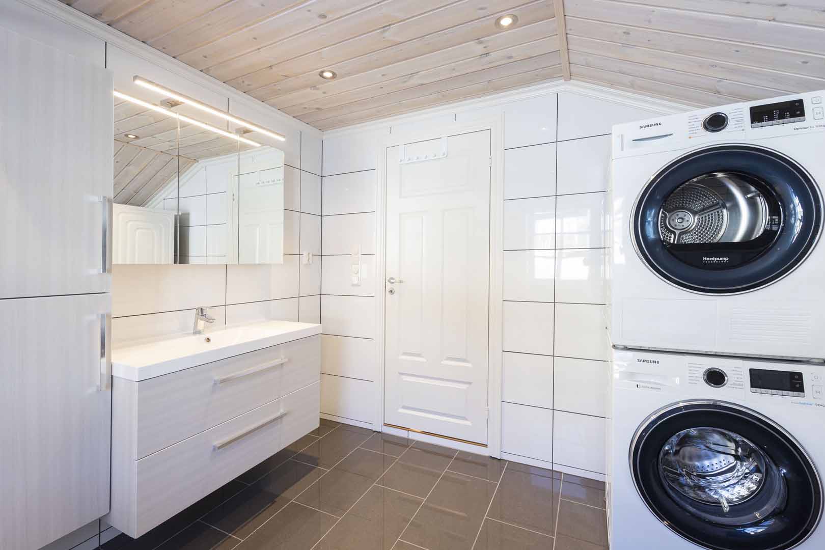Moderne vaskerom med vaskemaskin og tørketrommel, hvite fliser og lys trepanel i taket.