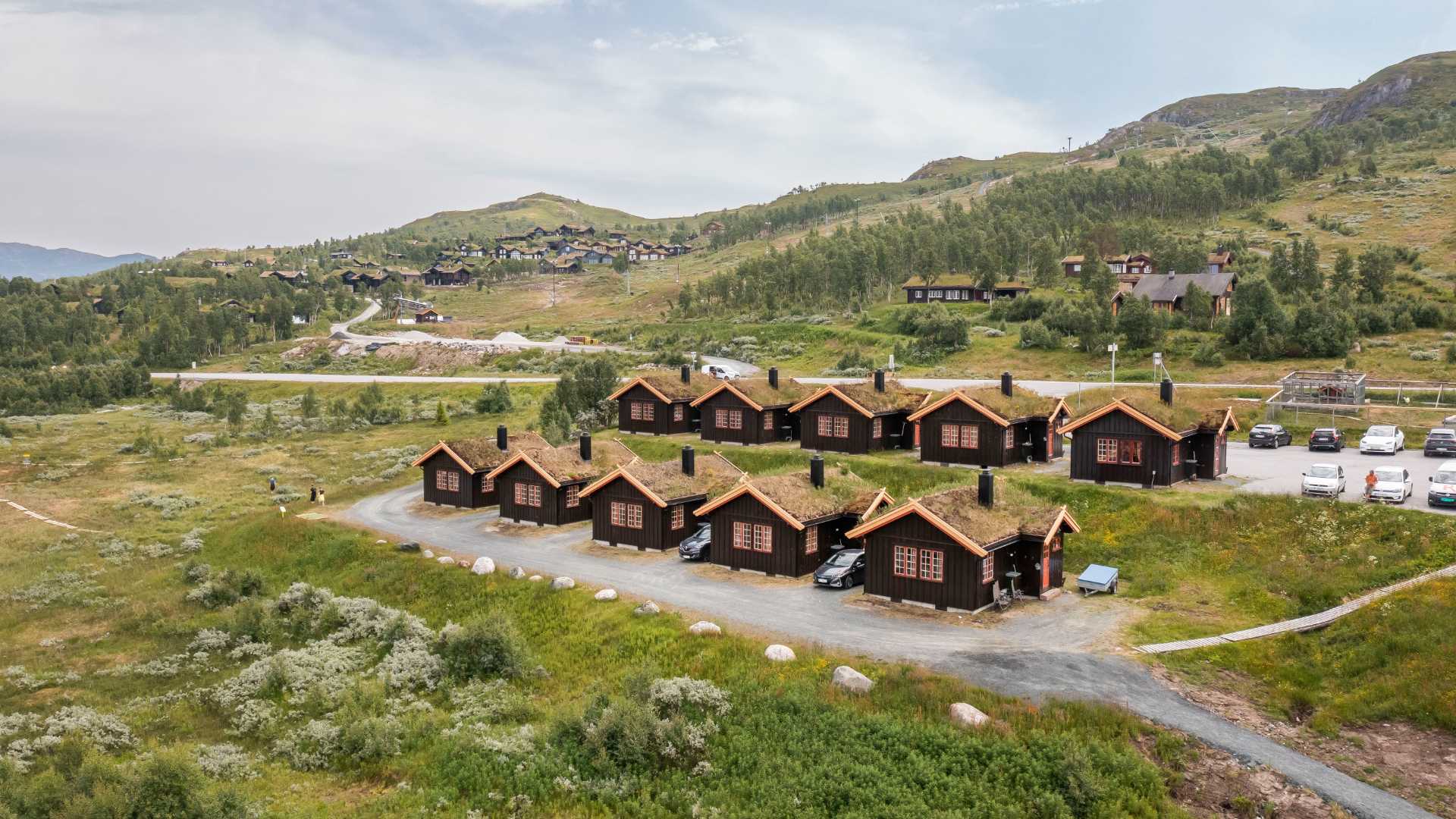 Hyggelige norske tømmerhytter i fjellet med gress på takene, omgitt av grønt landskap og bilparkering.