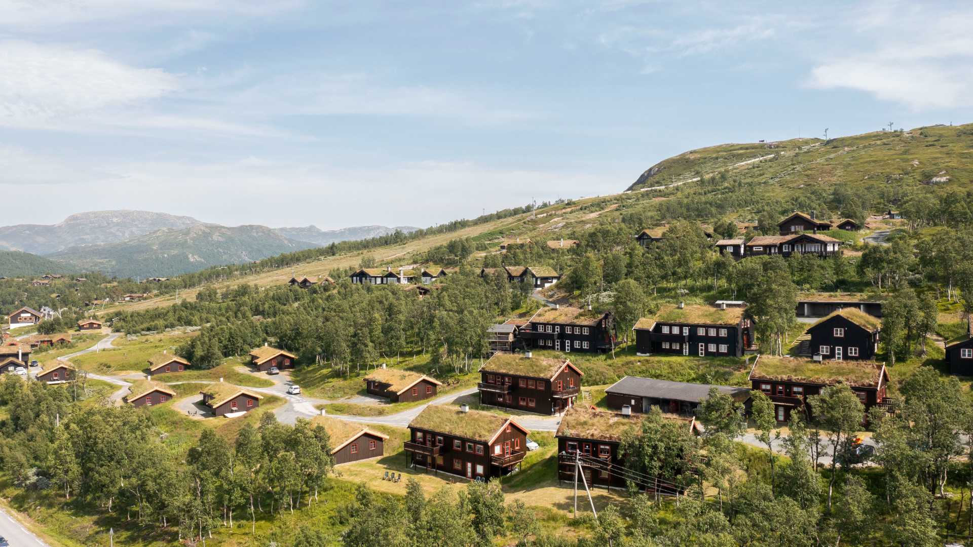 Norsk fjellandskap med tradisjonelle trehytter og grønne tak, omgitt av skog og fjell.