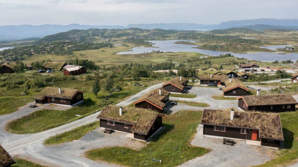 Landskap med flere hytter med torvtak, innsjø i bakgrunnen og grønn natur, sett fra høyden.