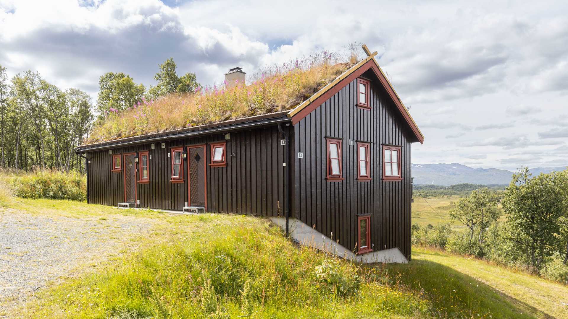 Svart, tradisjonell norsk hytte med torvtak i naturskjønt landskap med fjell i bakgrunnen under overskyet himmel.