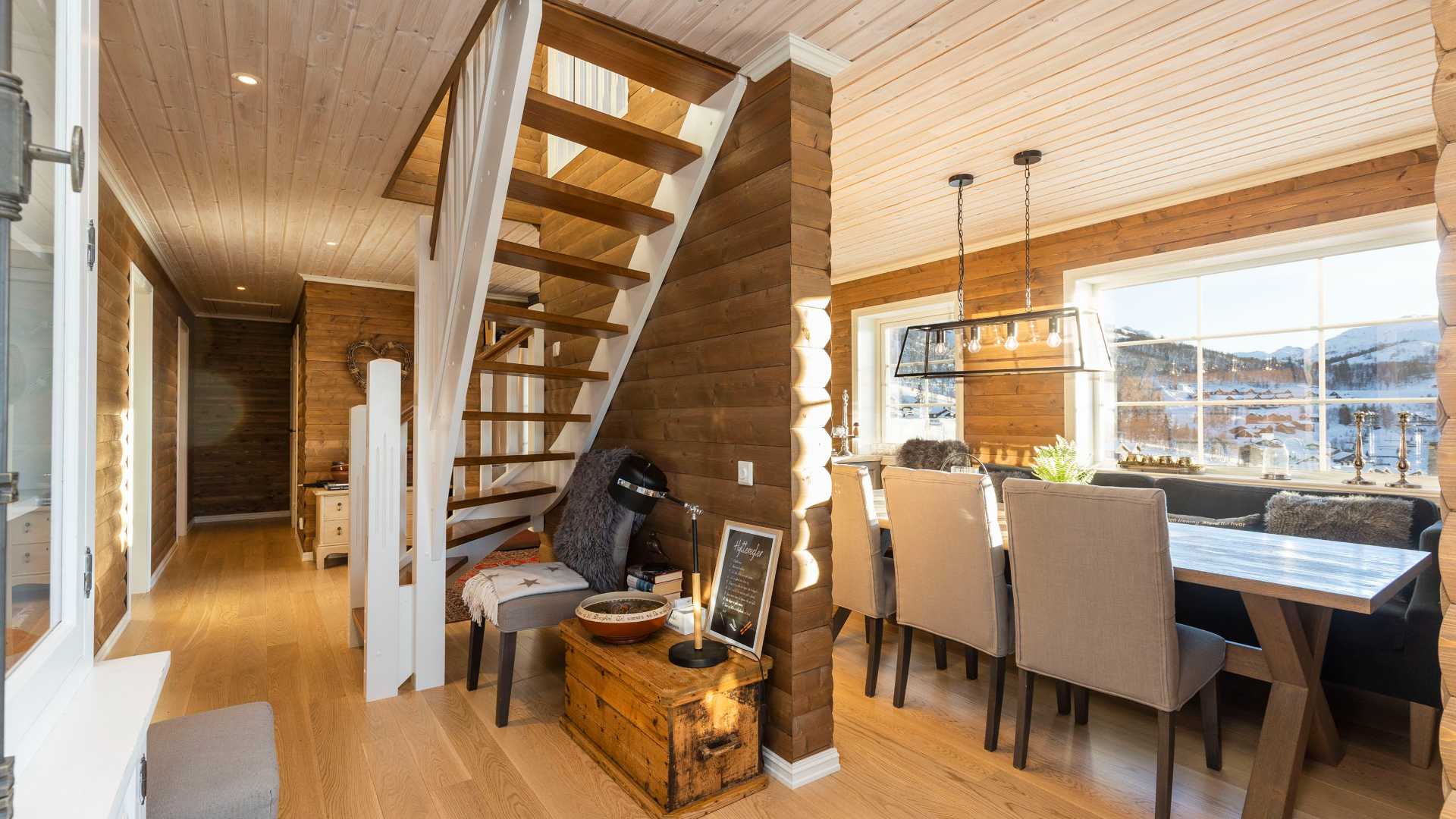 Moderne, koselig hytteinteriør med spisebord, åpne trapper og store vinduer med utsikt over snødekte fjell.