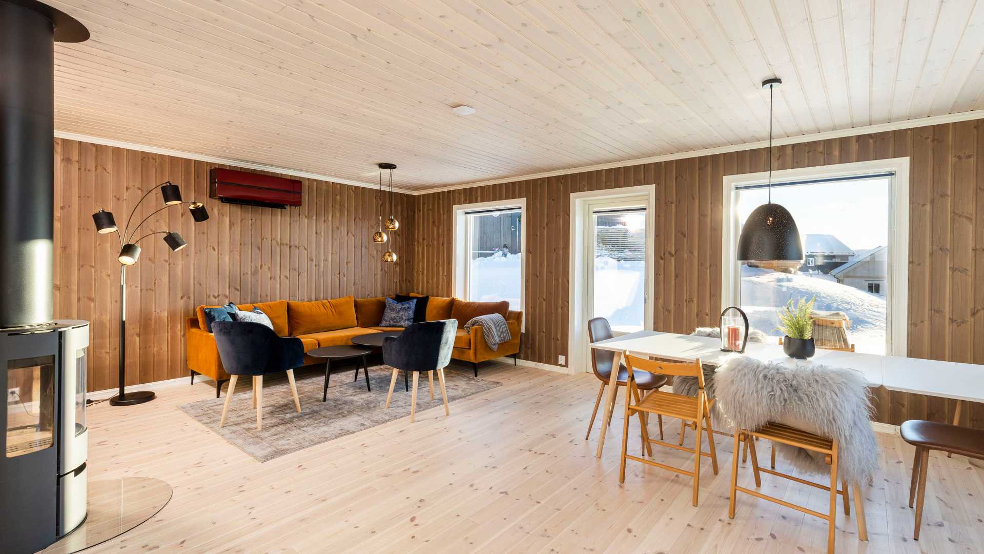 Moderne stue med trepanelvegger, oransje sofa og spisebord med utsikt mot snødekt landskap gjennom store vinduer.