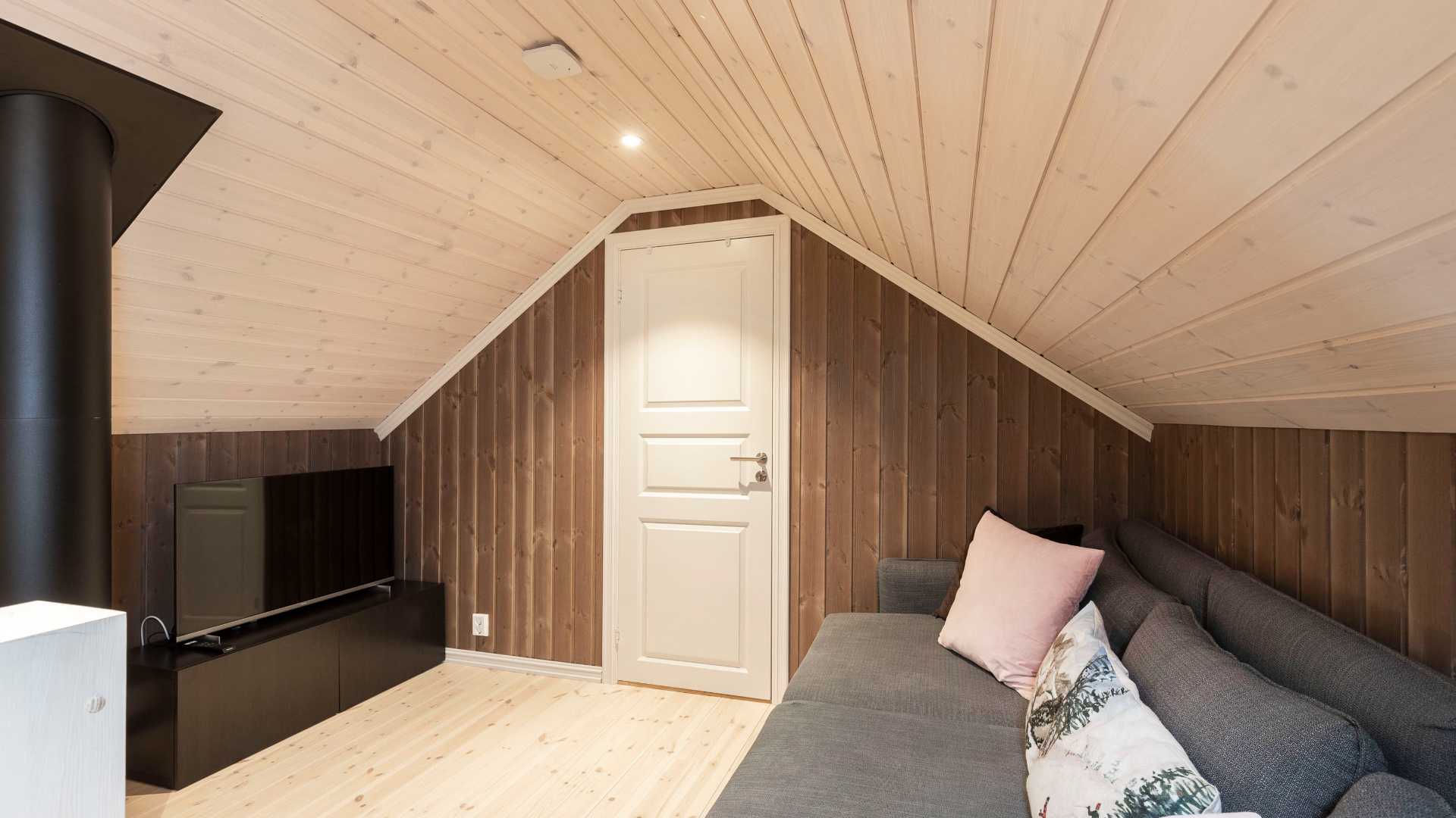 Loftstue med tregulv, komfortabel grå sofa og TV, vegger i trepanel og koselig interiør.