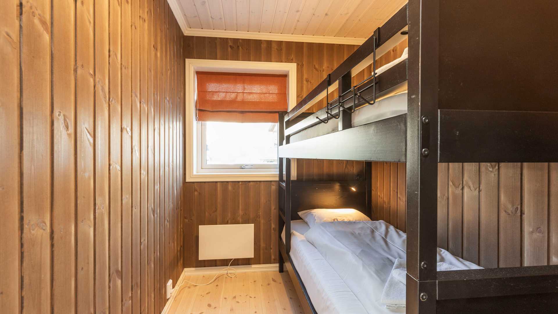 Smalt soverom med køyeseng, trepanelvegger og vindu med rød rullegardin. Koselig hytteinteriør.
