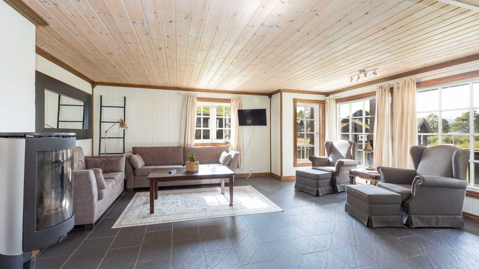 Lyst stueinteriør med sofa, stoler, peis og store vinduer som slipper inn mye naturlig lys.