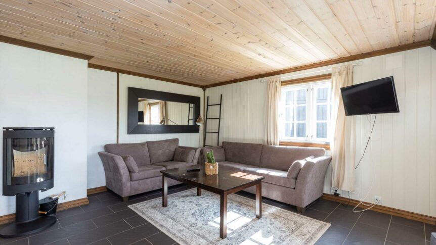 Stue med peis, grå sofaer, TV på veggen og et stort speil, lyst interiør og tregulv i en koselig hytte.