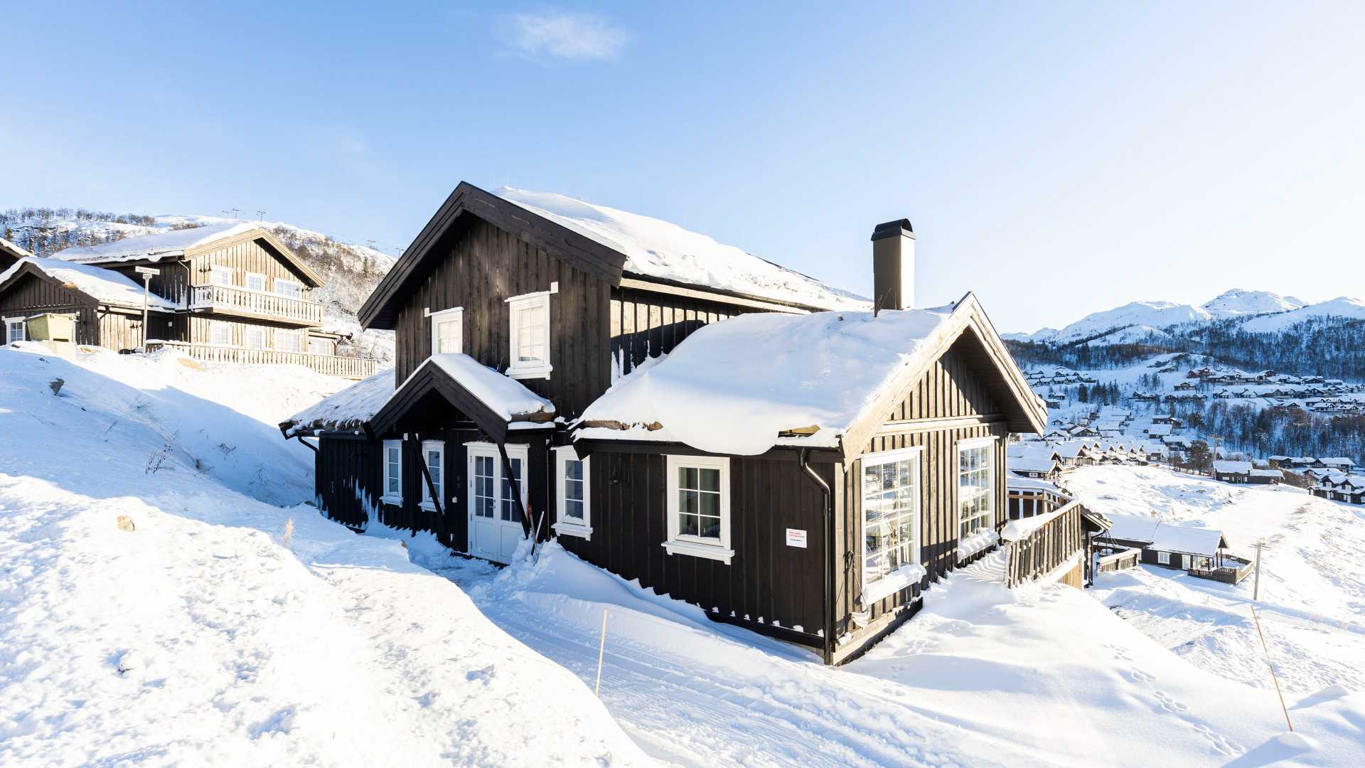 Tradisjonell norsk hytte dekket av snø i et naturskjønt vinterlandskap med fjell i bakgrunnen.