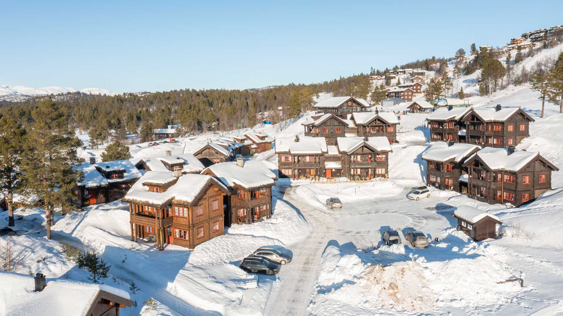 Snødekte hytter i fjellet, omgitt av vinterlig landskap og trær, under en klar blå himmel.
