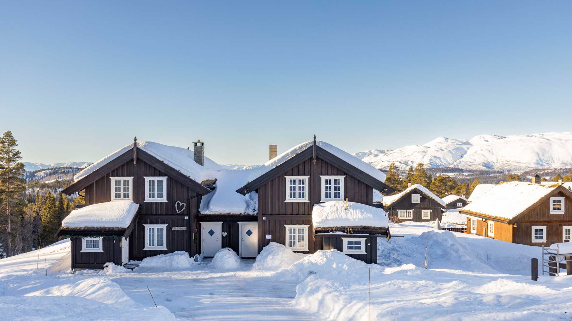 Snødekte hytter med fjellandskap i bakgrunnen på en solrik vinterdag i Norge.