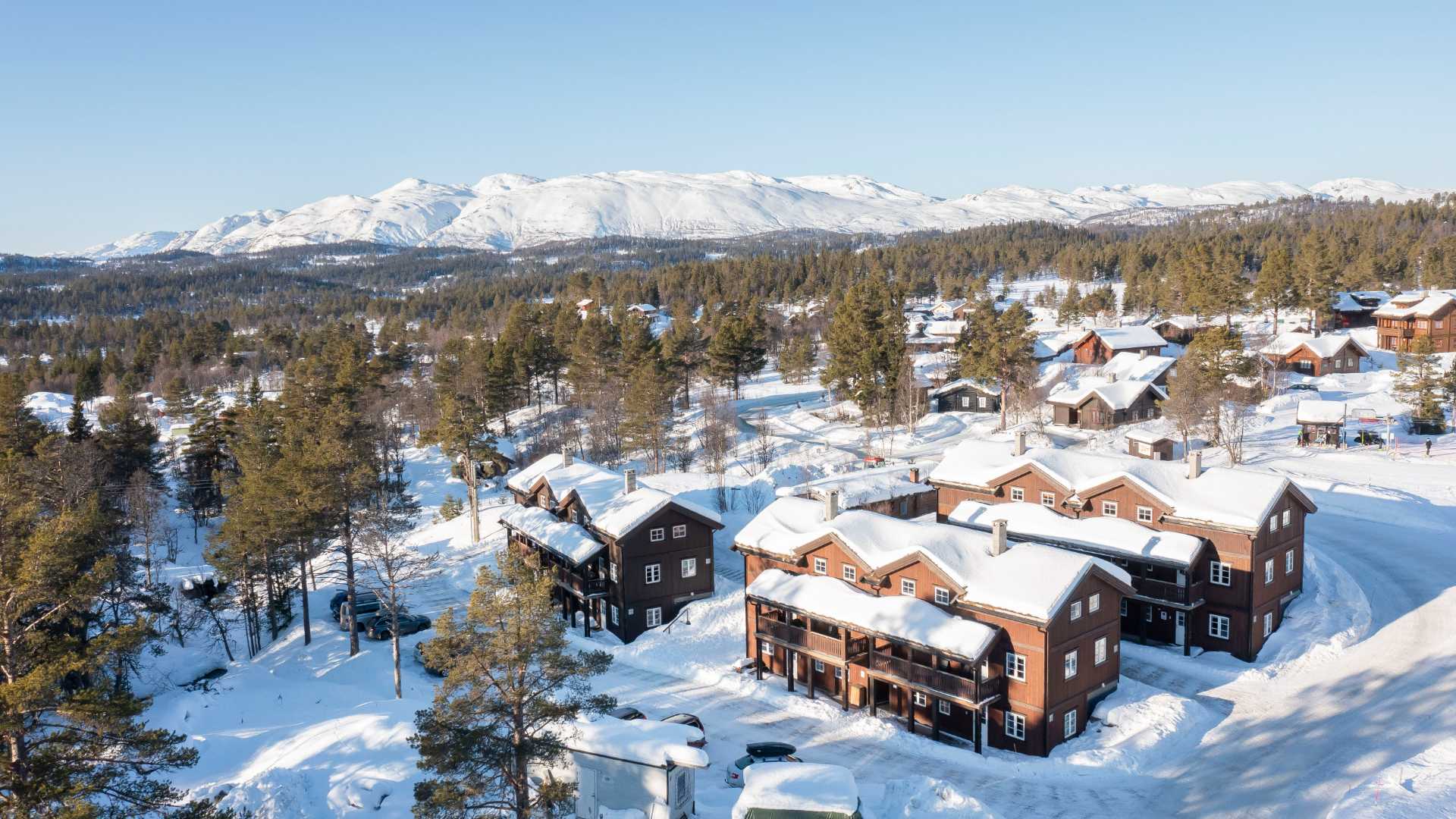 Snødekt norsk fjellandsby med trehus og skog i bakgrunnen, under klar blå himmel.
