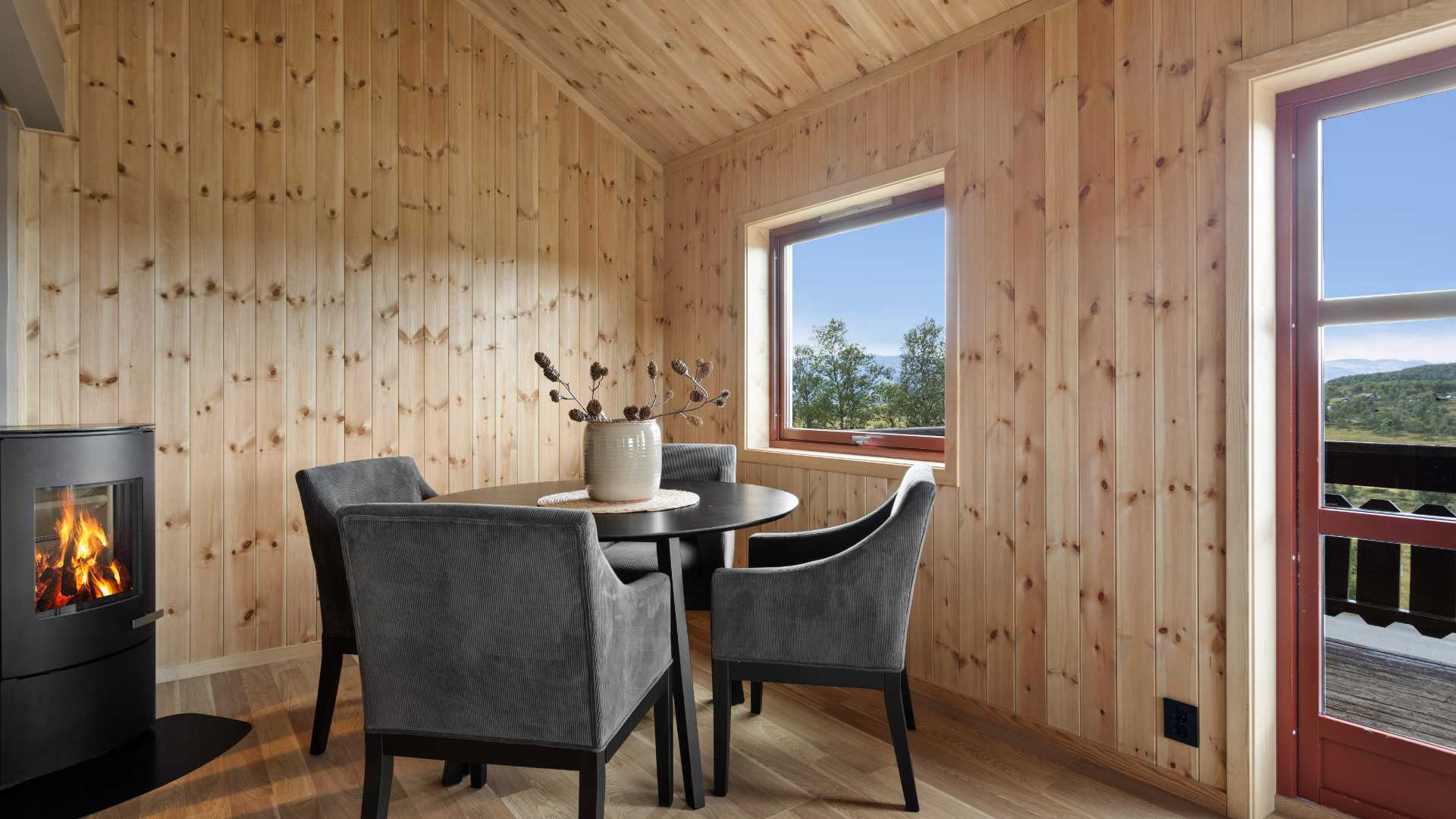Koselig hytteinteriør med peis, bord og stoler, utsikt til naturen gjennom vinduet.