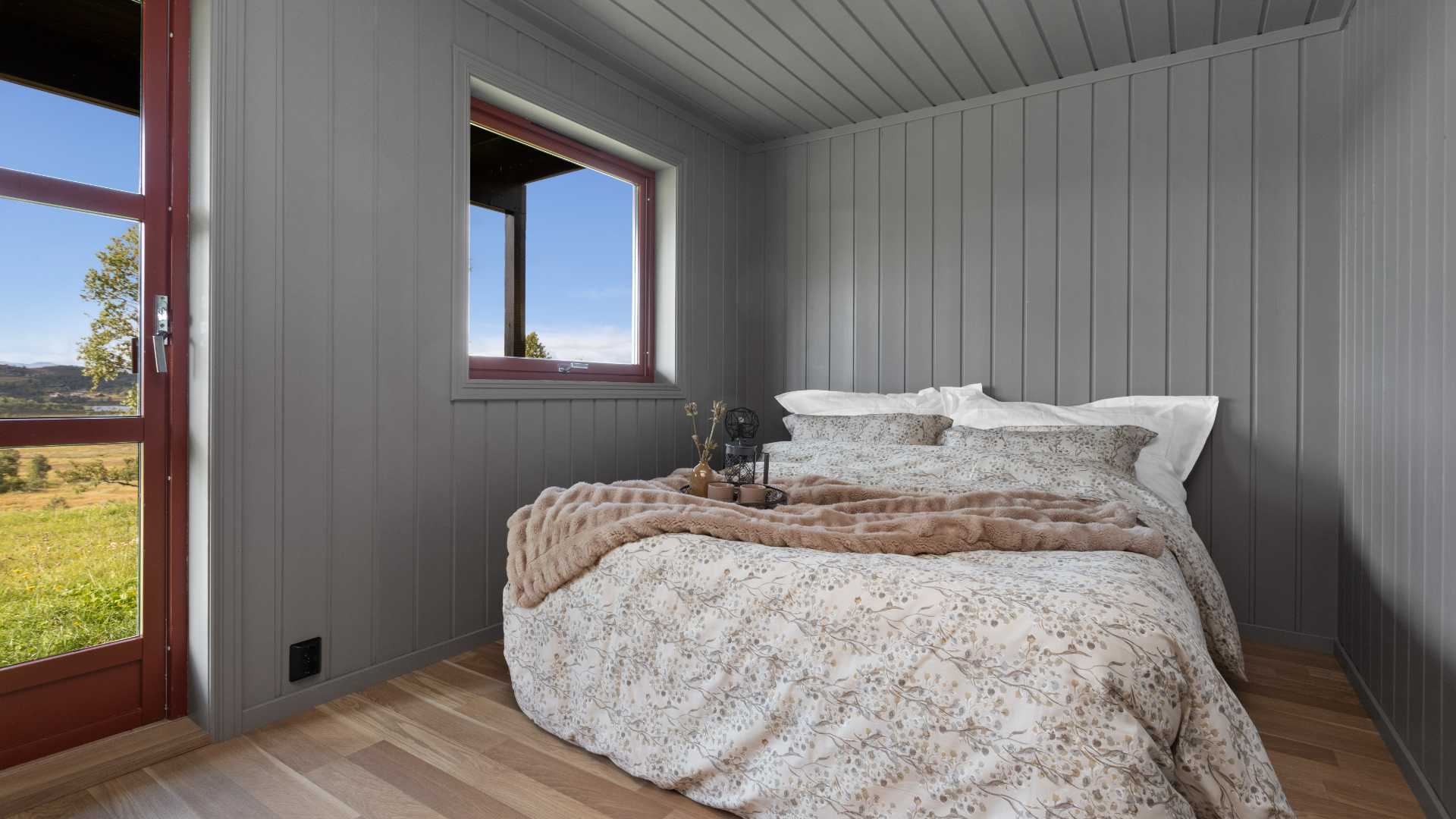 Koselig soverom med blomstret sengesett, grå vegger og vindu med utsikt til grønt landskap og blå himmel.