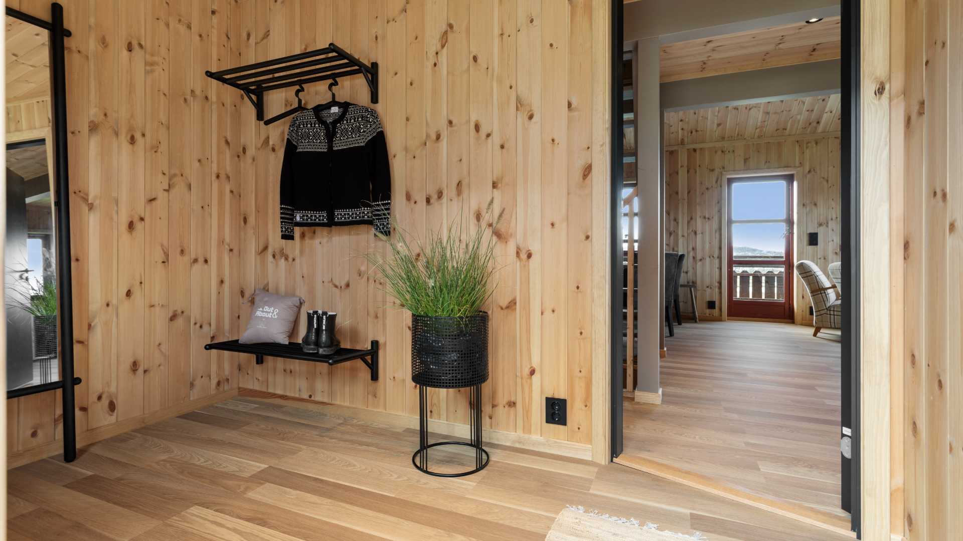 Gang med trepanel, klesstativ, svarte sko og plante, med utsikt til stue med store vinduer.