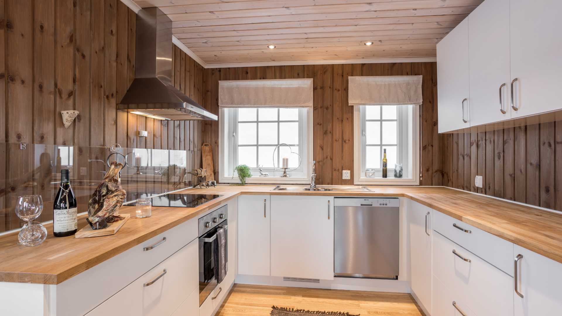 Moderne kjøkken med trepanel, lysinnslipp fra vinduer, hvite skap og benkeplate i tre.