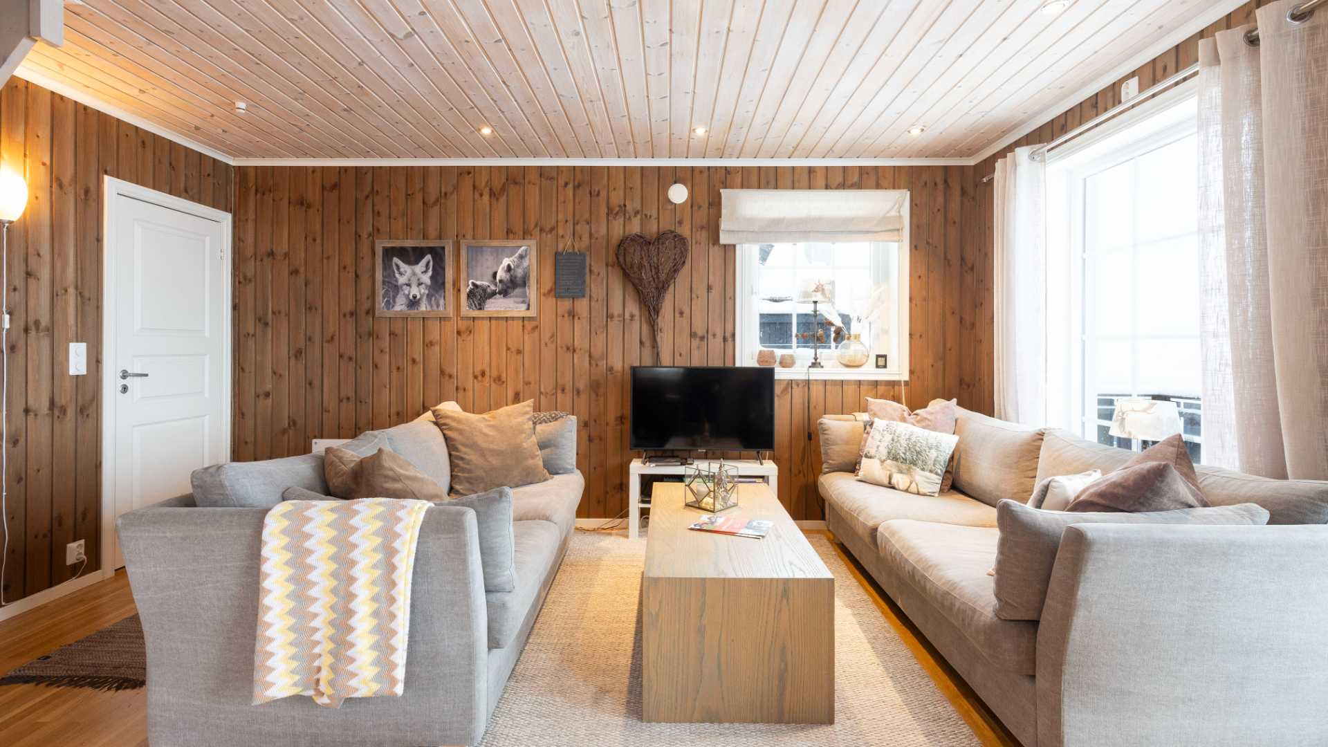 Moderne stue med hvitt tak, trepaneler, grå sofaer, TV og vindu med gardiner.