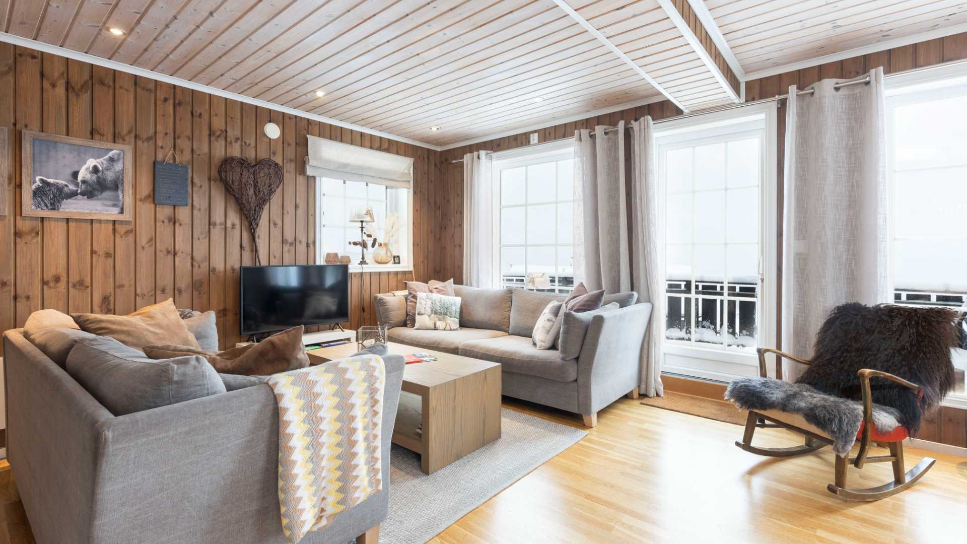 Stue med tregulv, grå sofa, skinntrukket gyngestol og store vinduer med snødekt utsikt.