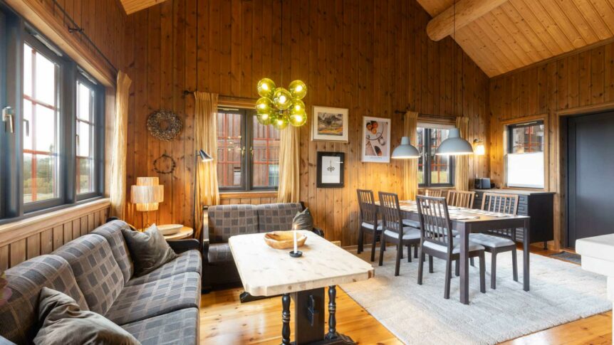 Koselig stue med tregulv, sofa, spisebord og moderne belysning i en hytte med store vinduer.