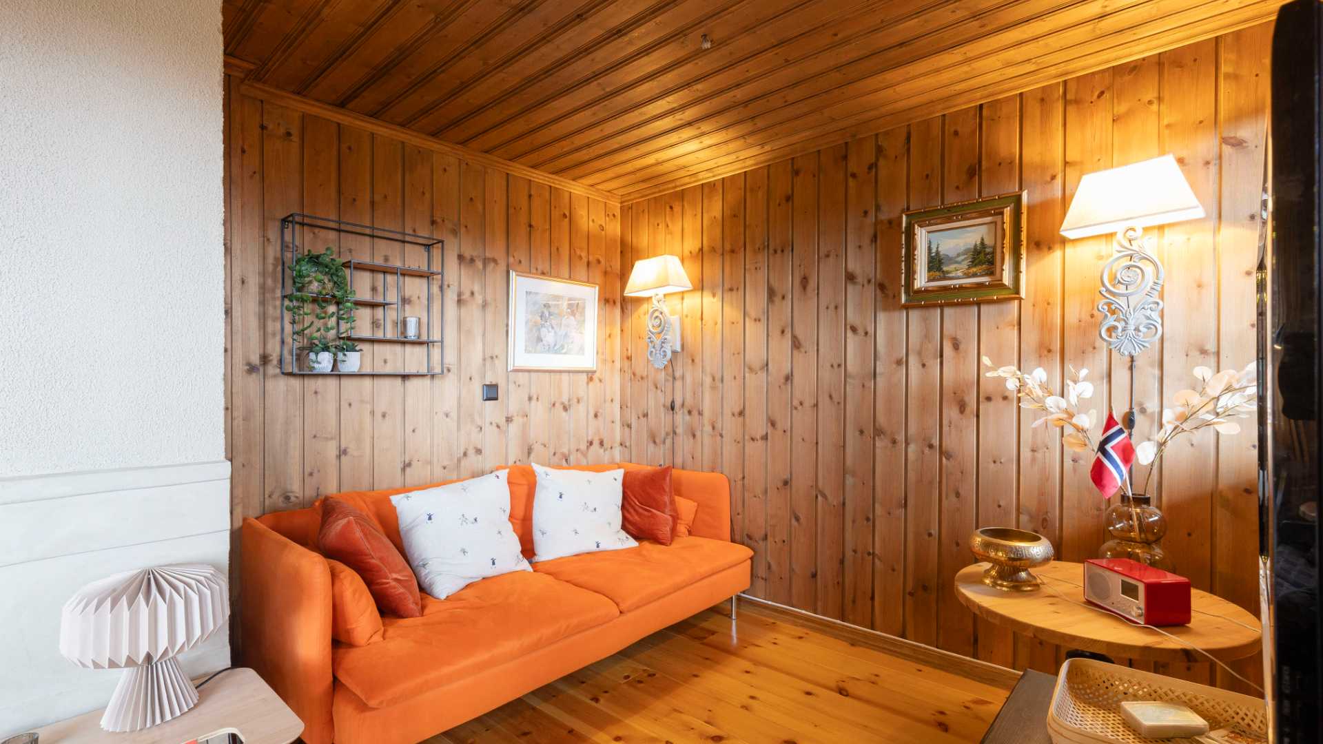 Koselig norsk stue med oransje sofa, trepaneler, kunstverk og norsk flagg på et trebord.