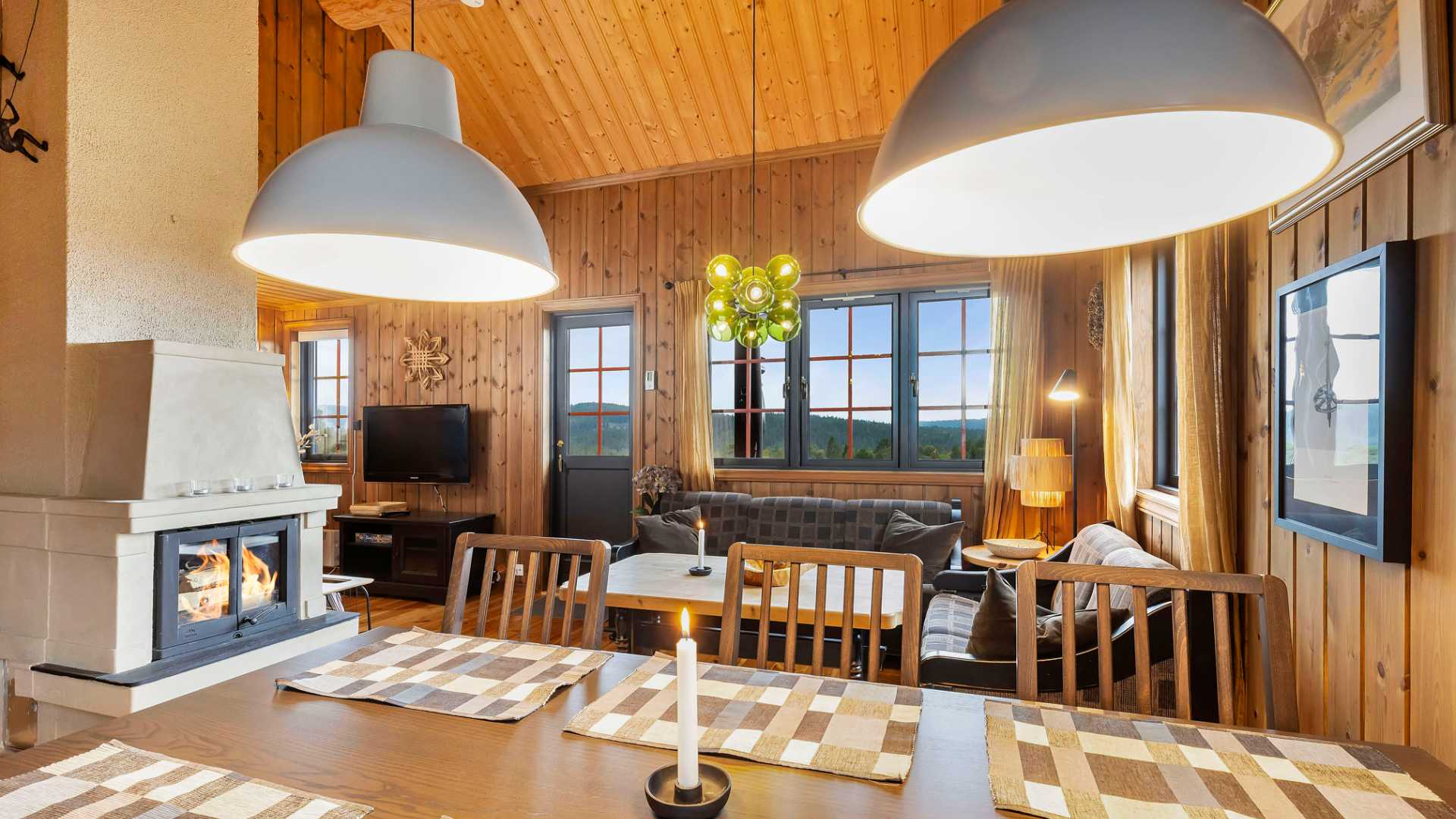 Koselig stue i tømmerhytte med peis, spisebord og store vinduer som gir utsikt til naturen utenfor.