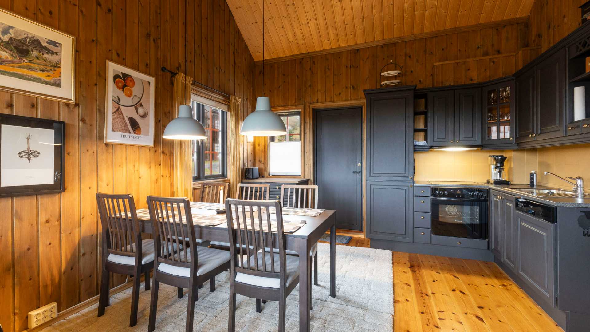 Koselig kjøkken- og spisestue med trepanel, mørke skap og spisebord for seks personer under moderne hengelamper.