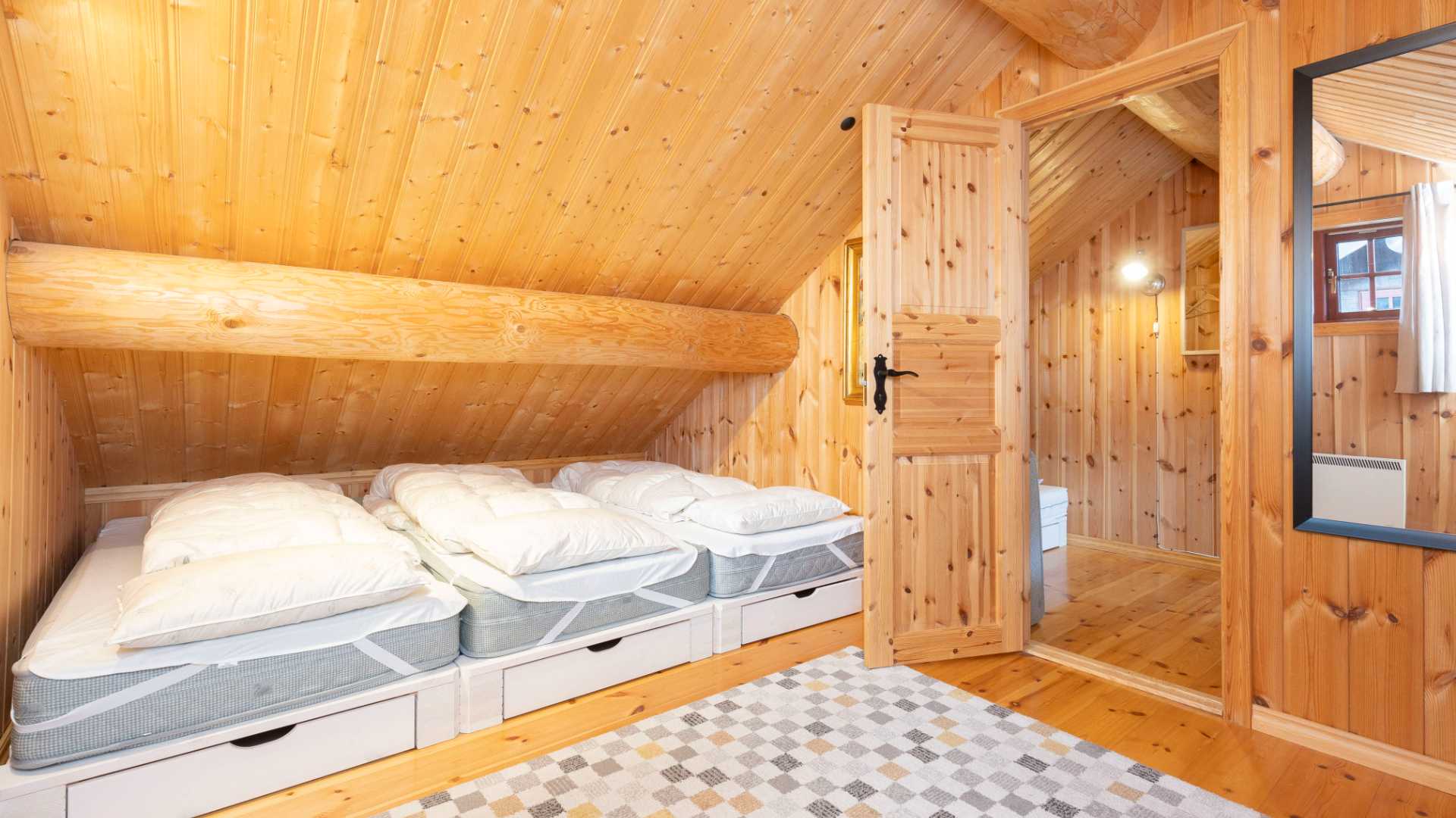 Loftsrom i tre med tre senger under skråtak, naturlig lys fra vindu, inngang til et annet rom.