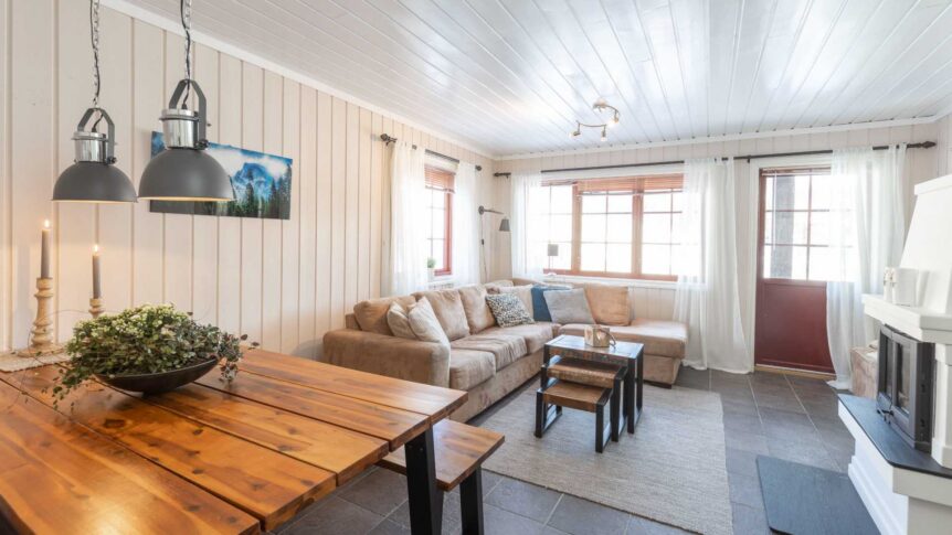 Moderne stue med beige sofa, trebord, lys innredning og store vinduer med gardiner.