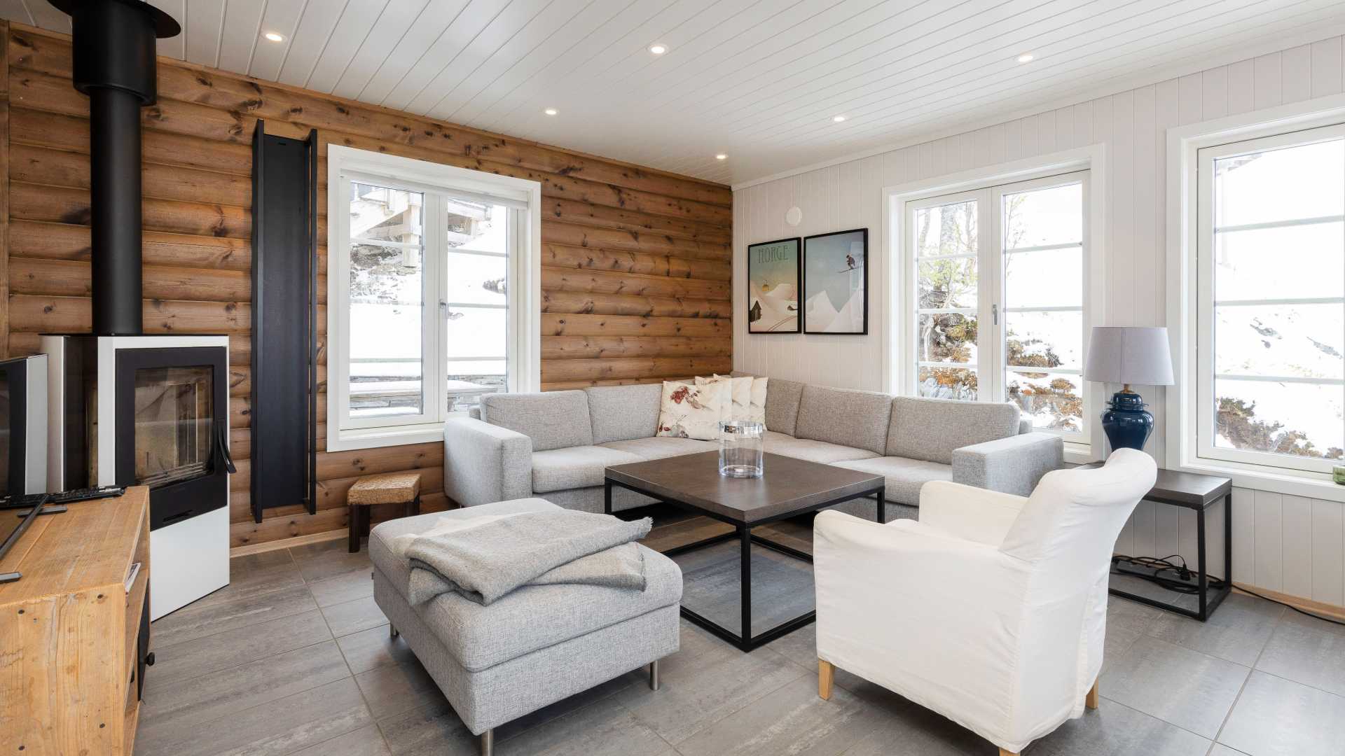 Moderne stue med peis, grå sofaer, og lyse vegger. Store vinduer slipper inn naturlig lys. Koselig norsk interiør.