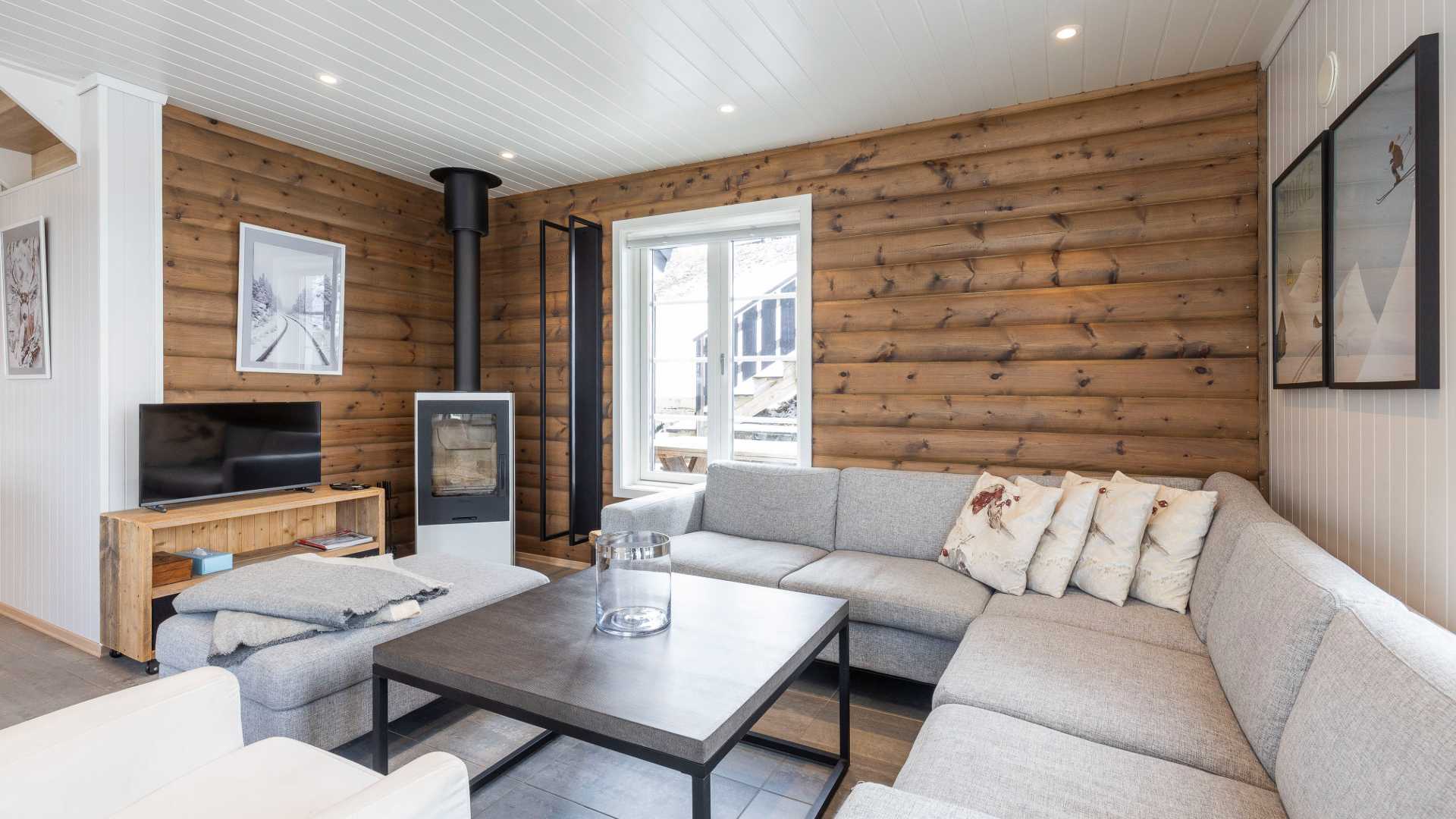 Stue med grå sofa, peis, og trekledning på veggen. Hyggelig og moderne hytteinteriør med bilder og TV.