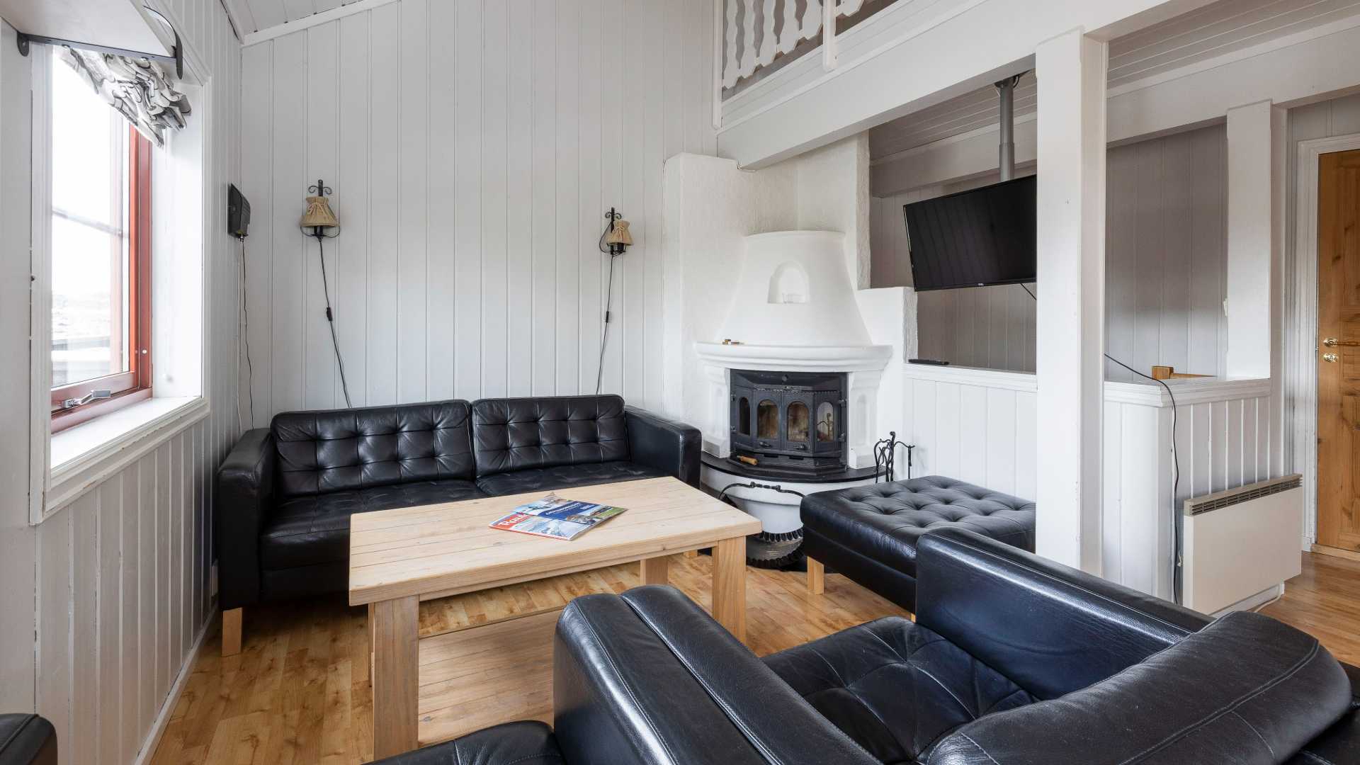 Stue med svart skinnsofa, trebord, peis og TV på veggen i et lyst, moderne rom.