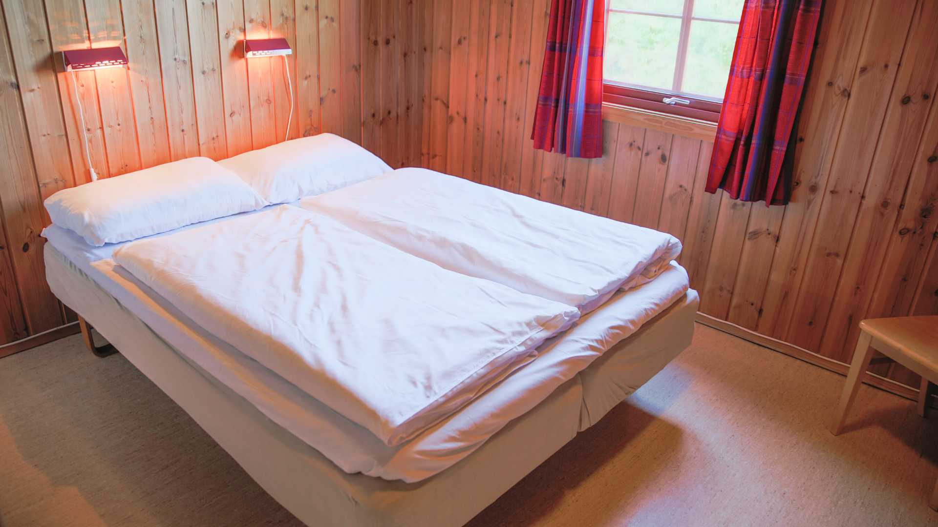 Seng med hvitt sengetøy i en koselig hytte med trepanel, røde gardiner og nattlamper.