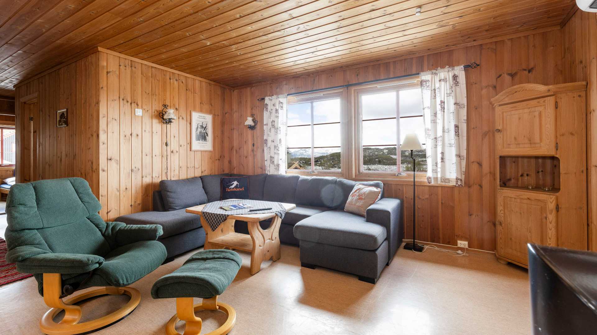 Koselig stue med trepanel, hjørnesofa, lenestol og store vinduer med utsikt over landskapet.