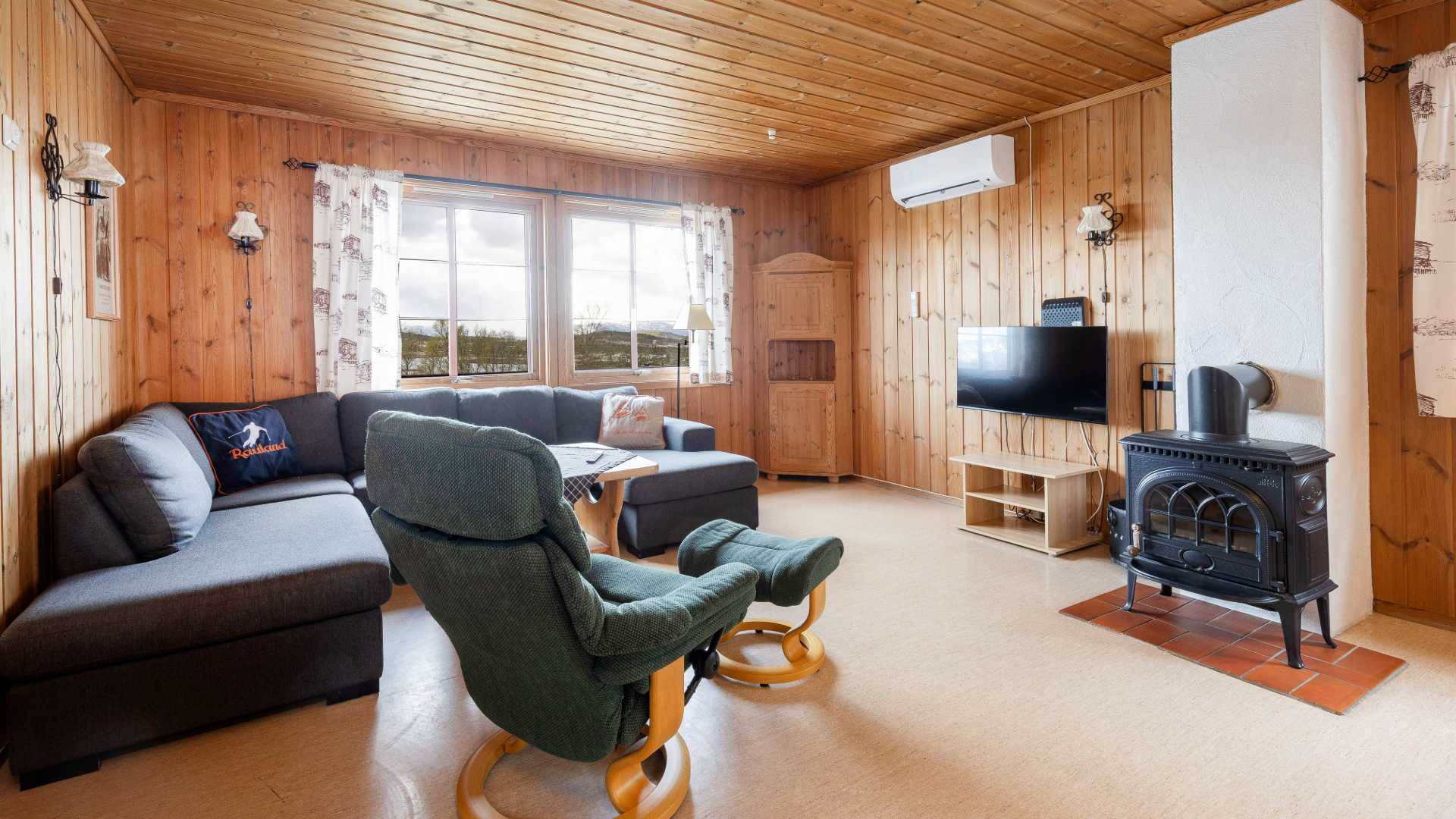 Koselig norsk stue med tregulv, peis, TV, og komfortable sitteplasser. Store vinduer gir naturlig lys.