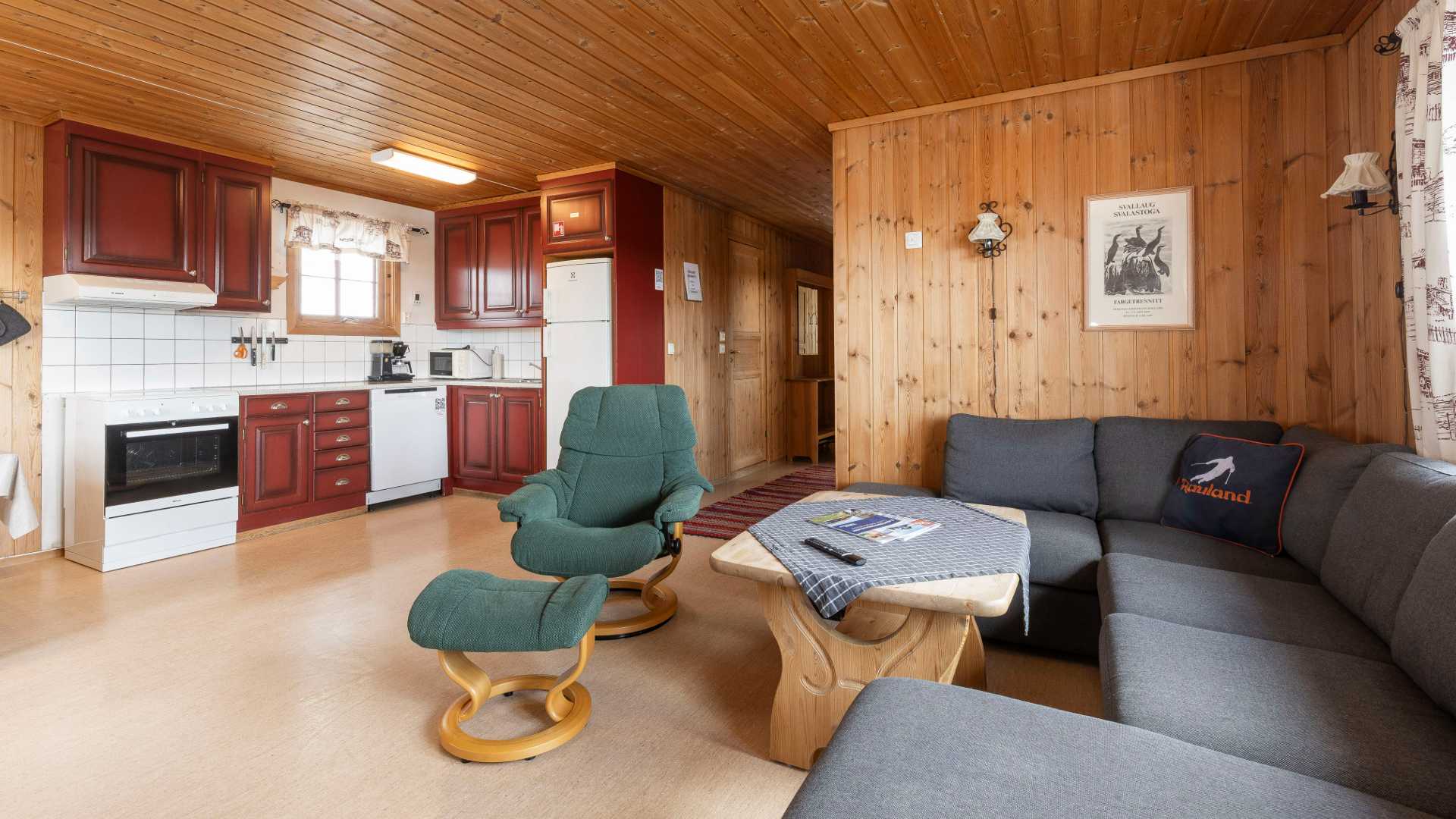 Koselig hyttestue med trepanel, peis, kjøkkenkrok og grå sofa. Perfekt for avslapning.