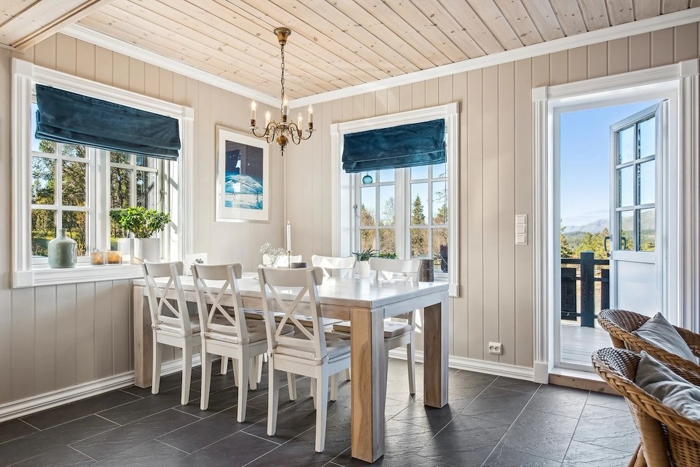 Lyst spisestue med trebord, hvite stoler, lysekrone og utsikt til naturen gjennom store vinduer. Skandinavisk stil.