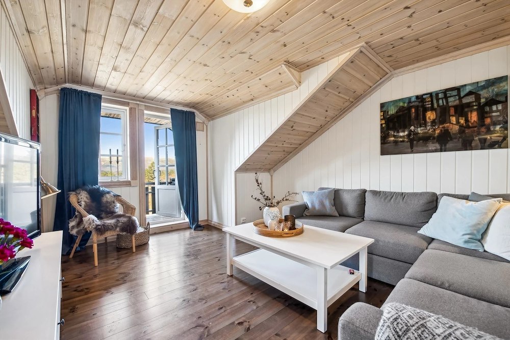Lys og koselig stue med tregulv, grå sofa, hvitt bord og kunst på veggen, dekorert med blå gardiner og ullpute.