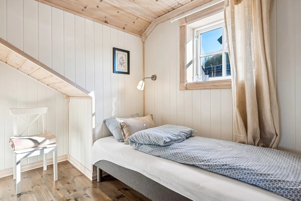 Lyst og koselig soverom med enkeltseng, trepanelvegger og et lite vindu med gardin. En stol i hjørnet.
