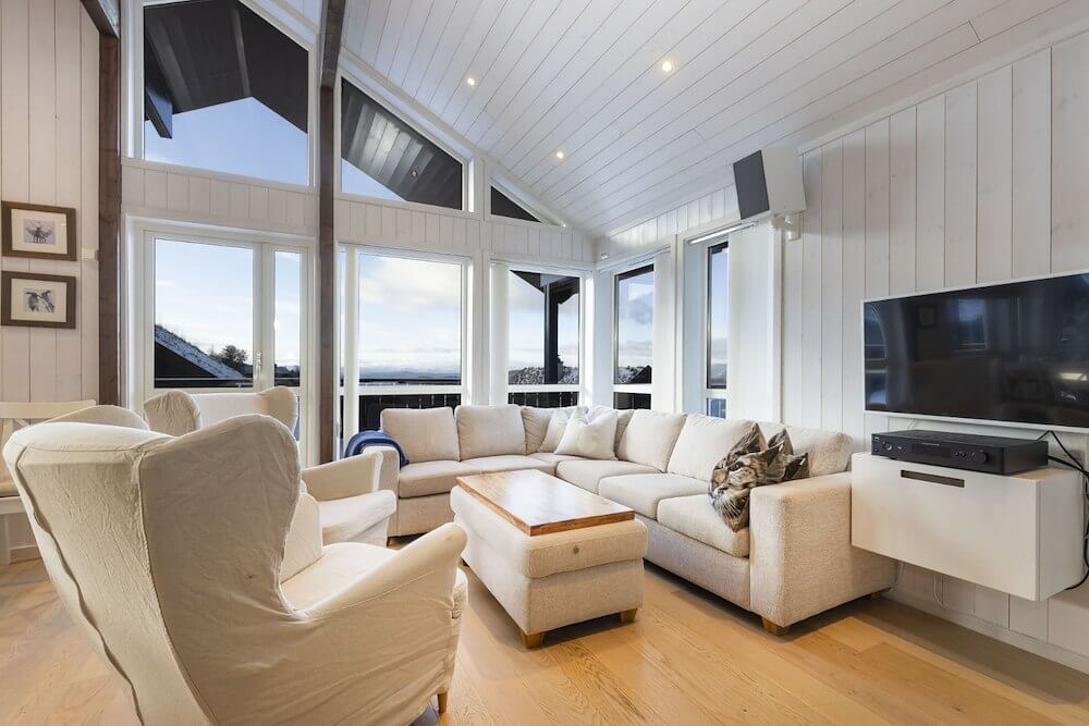 Lys og romslig stue med moderne innredning, stor sofa, TV og panoramavinduer med utsikt.
